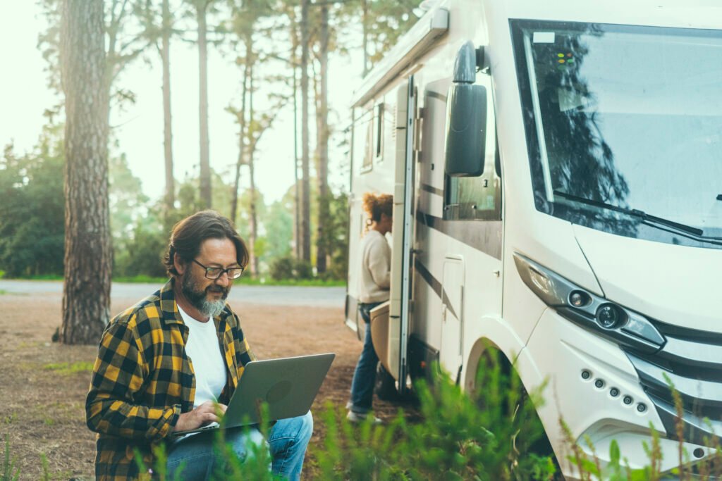 Man using a laptop beside an RV at a forest campsite, checking RV systems during a road trip