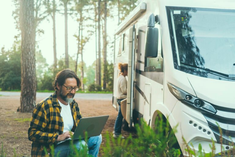 Man using a laptop beside an RV at a forest campsite, checking RV systems during a road trip